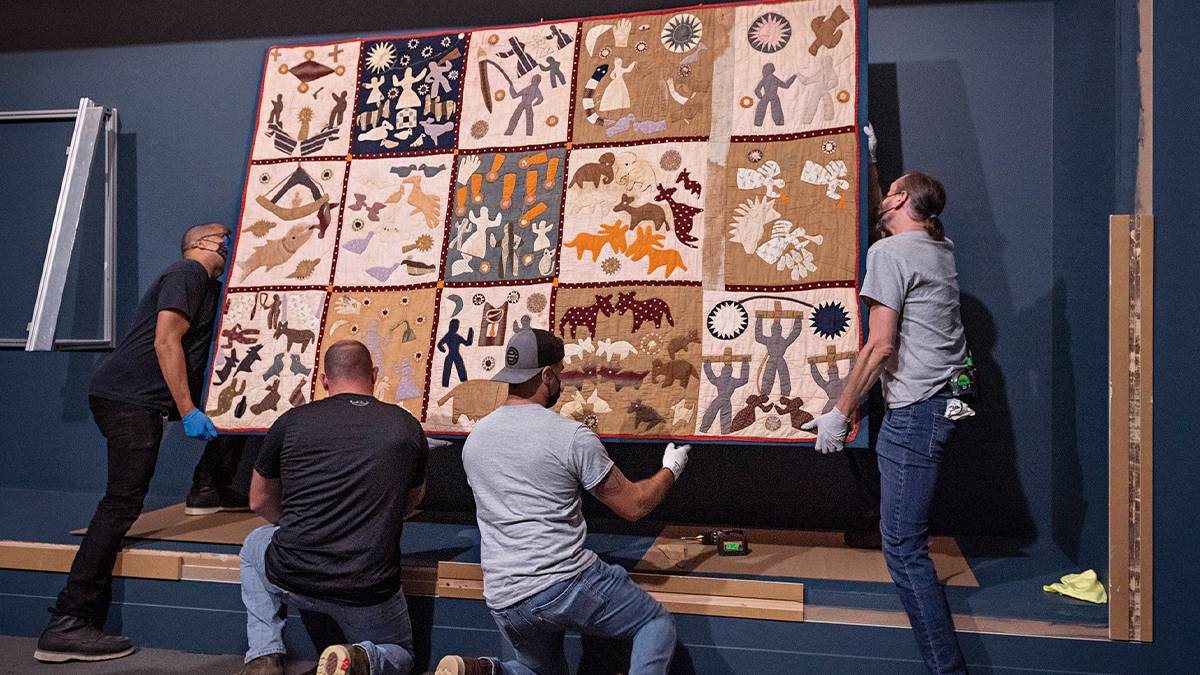 Four men in the process of hanging an elaborate quilt on a blue wall in Boston, Massachusetts