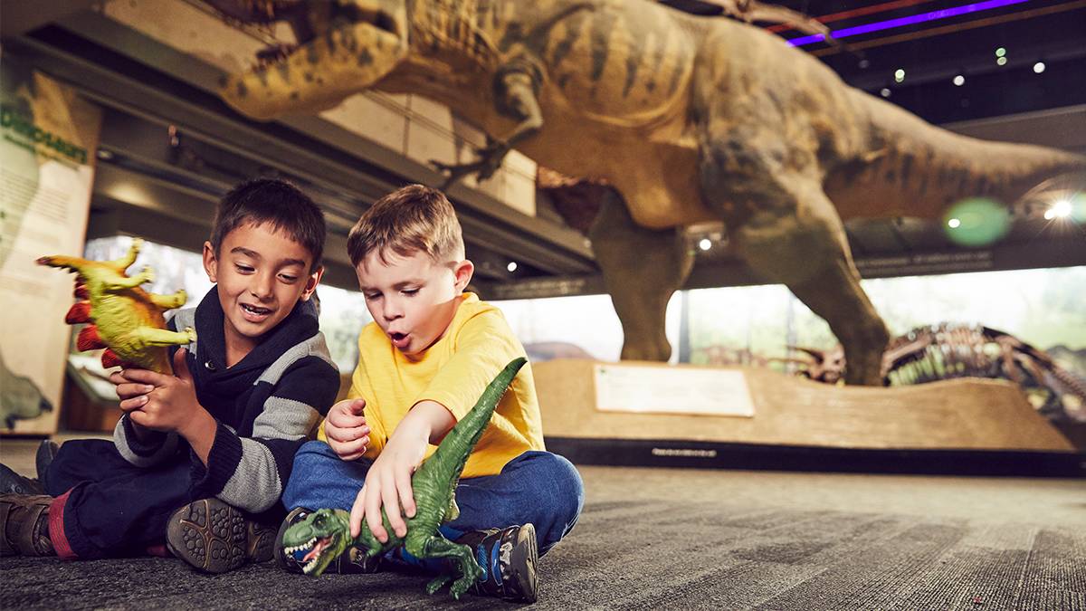Two kids playing with toy dinosaurs with a life size t-rex in the background at the Museum of Science in Boston, Massachusetts