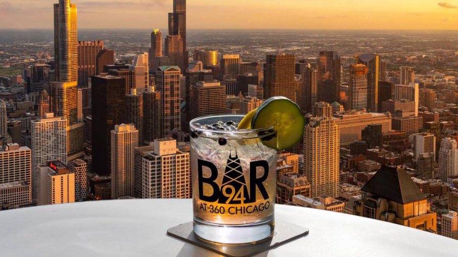 A mixed drink in a clear glass with a cucumber garnish on a table overlooking Chicago at Sunset