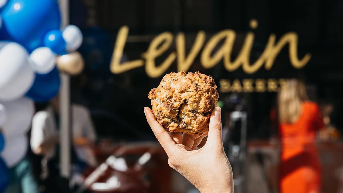 Close up shot of a hand holding a muffin with the wind sign for Levain Bakery in the background in NYC, New York