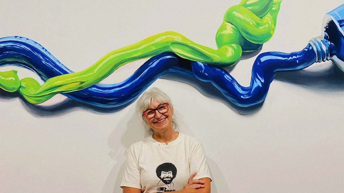 Tracy Lee Strum standing in front of one of her paintings of blue and green paint and large paint tubes at the TiLT Museum in Rutherford, New Jersey, USA