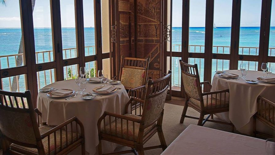 A dining room with several tall thin windows and two tables with white cloths and wooden chairs at La Mer in Honolulu, Hawaii, USA