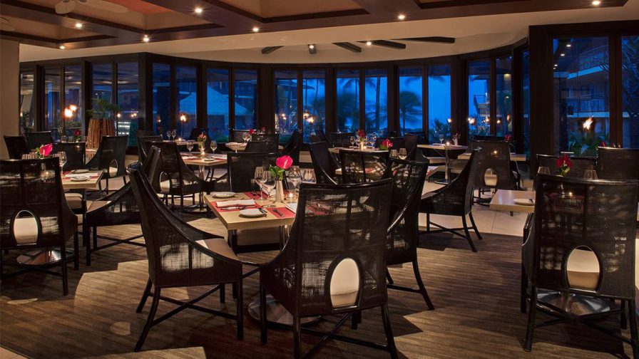 A large dining rom filled with dark chairs and lighter wooden tables with flowers on them and large window in the back showing nightfall at Red Salt in Kauai, Hawaii, USA