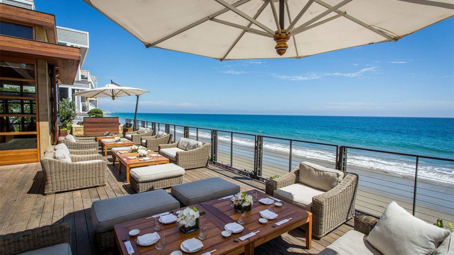 Out door dining area with wicker chairs, wooden tables, and big umbrellas with the ocean behind them at Nobu in Los Angeles, California,
