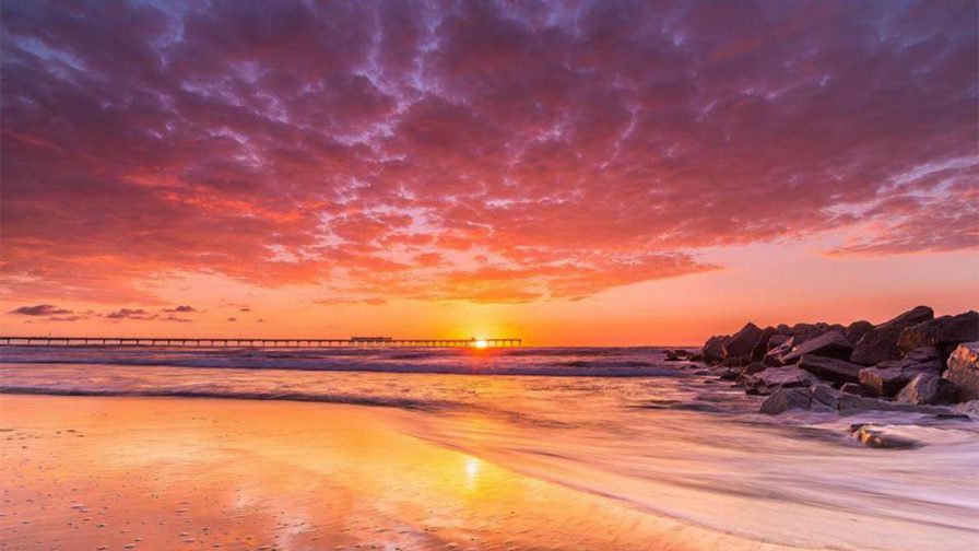 A bright orange, pink, and purple sunset with the Ocean Beach Pier in the background and rocks on the right hand sign in San Diego, California, USA