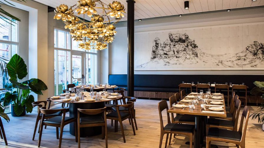 A dining room with large windows on the left and a black and white mural on the back wall with wooden tables and chairs plus a large gold floral chandelier at Mister Jiu’s in San Francisco, California, USA