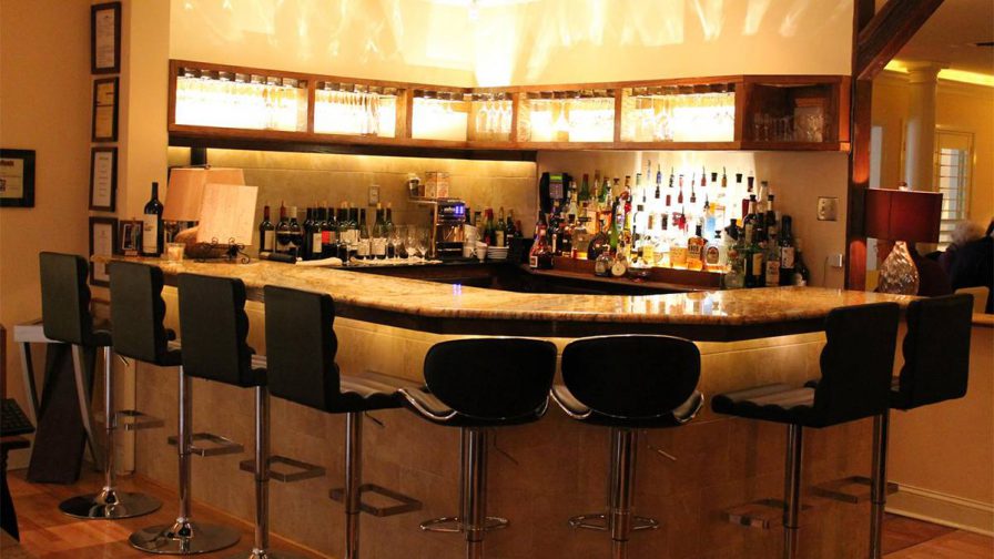 Close up photo of the bar at Le Yaca Restaurant with black seats and lights behind the wine glasses in the cabinet on the wall in Williamsburg, Virginia, USA