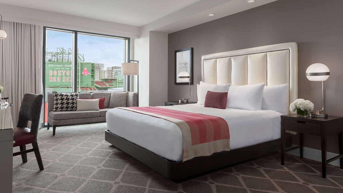 room at Hotel Commonwealth with grey carpeted floor and bed with white sheets and pink accents and white headboard, side tables and lamps, sofas, window with bilboard outside in Boston, Massachusetts, USA