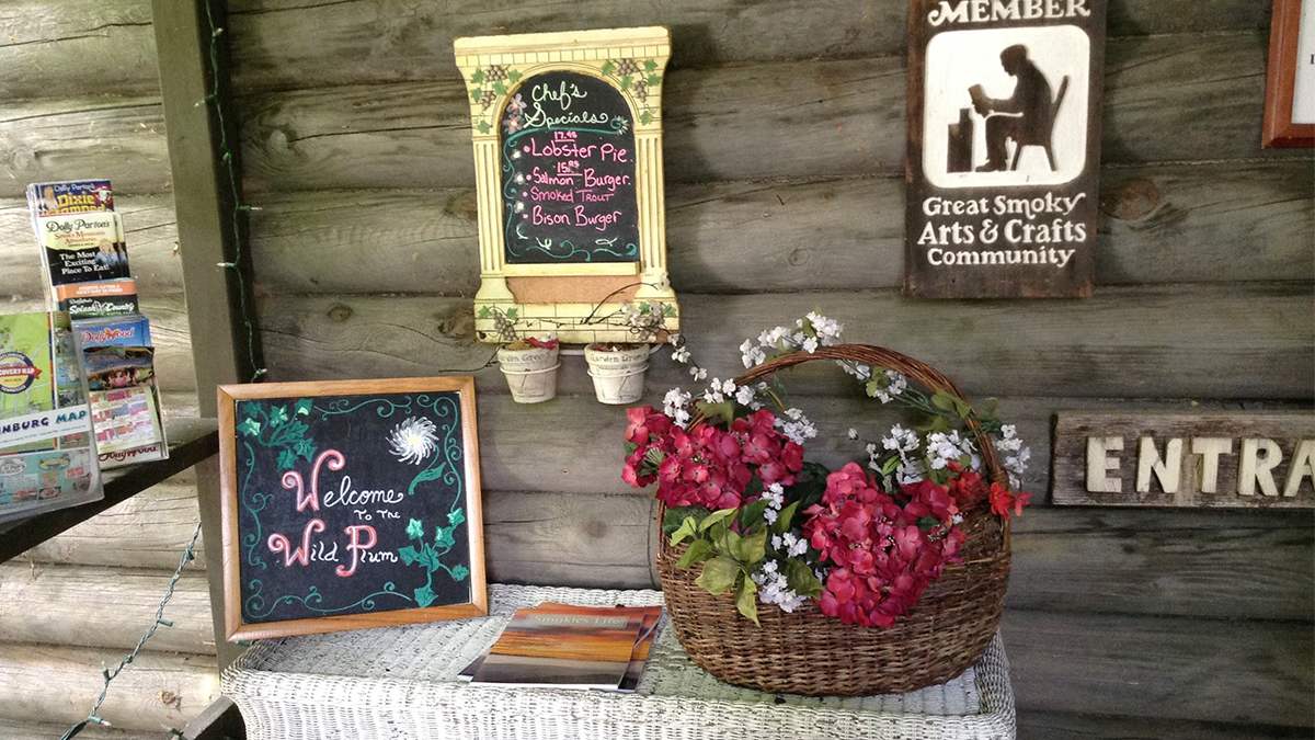 View of the entrance to Wild Plum Tea Room, a small white wicker tables with a welcome sign and a basket of flowers and a log wall behind it with a chef's specials menu on it in Gatlinburg, Tennessee, USA