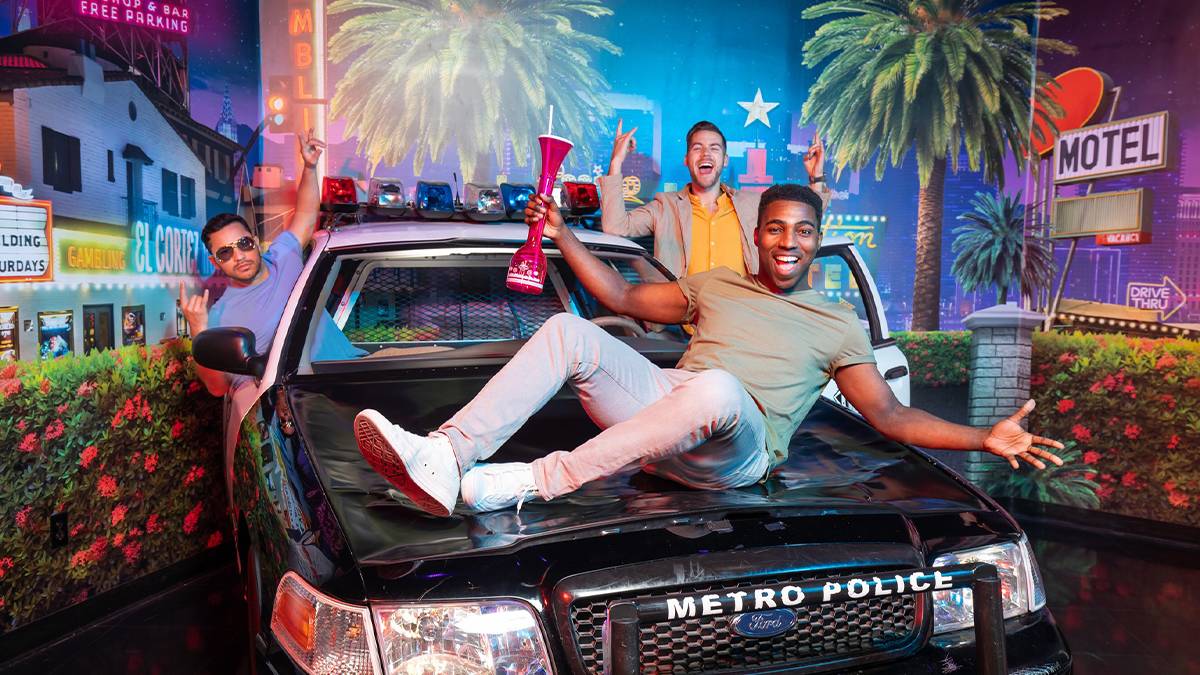 3 men posing with a police car one sitting on the good with a large drink in his hand and a mural of Vegas behind them at the The Hangover Bar Experience at Madame Tussauds in Las Vegas, Nevada, USA