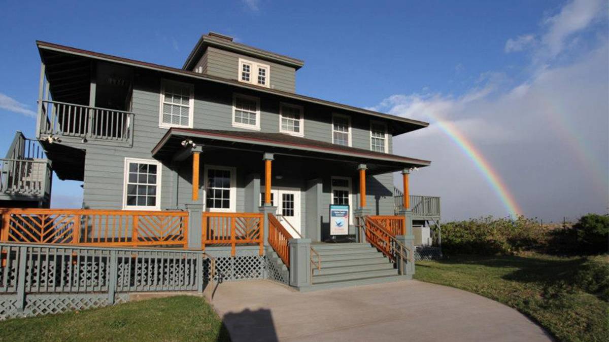 Ground view of the Hawaiian Islands Humpback Whale National Marine Sanctuary, a large muted green house with a wrap around porch and a deck off of the left side with two rainbows in the background in Maui, Hawaii, USA