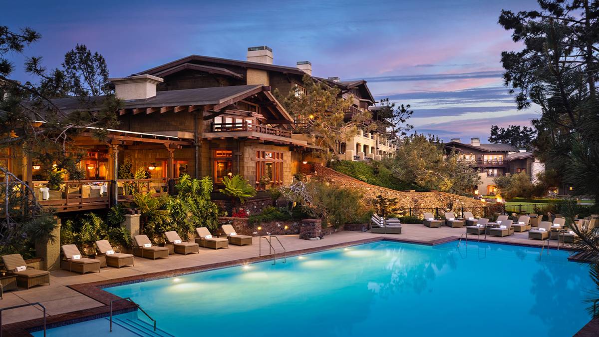 View of the Torrey Pine Lodge, a large multi level wooden and stone building with an outdoor dining area and it's pool lounge area with a light purple sunset in the background in San Diego, California, USA