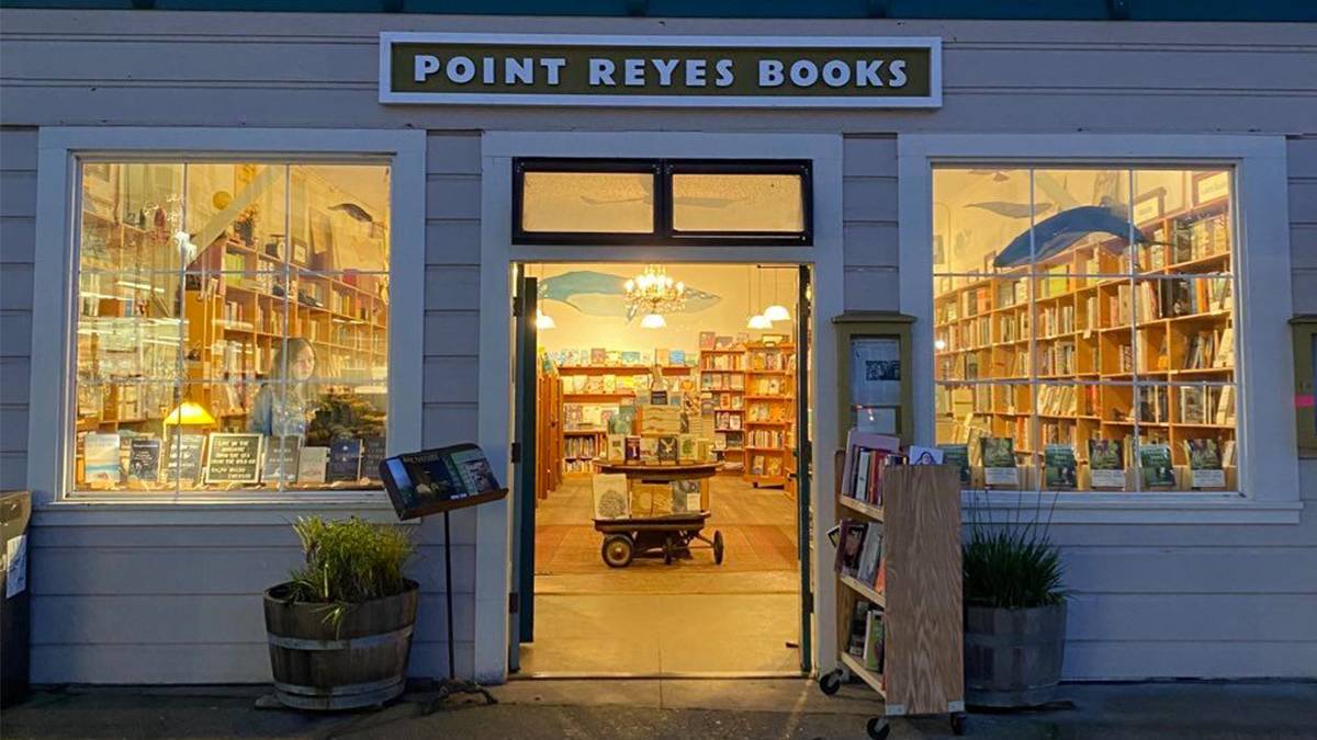 View of the front of Point Reyes Books at dusk with their lights on so you can see the store full of books and someone shopping in San Francisco, California, USA