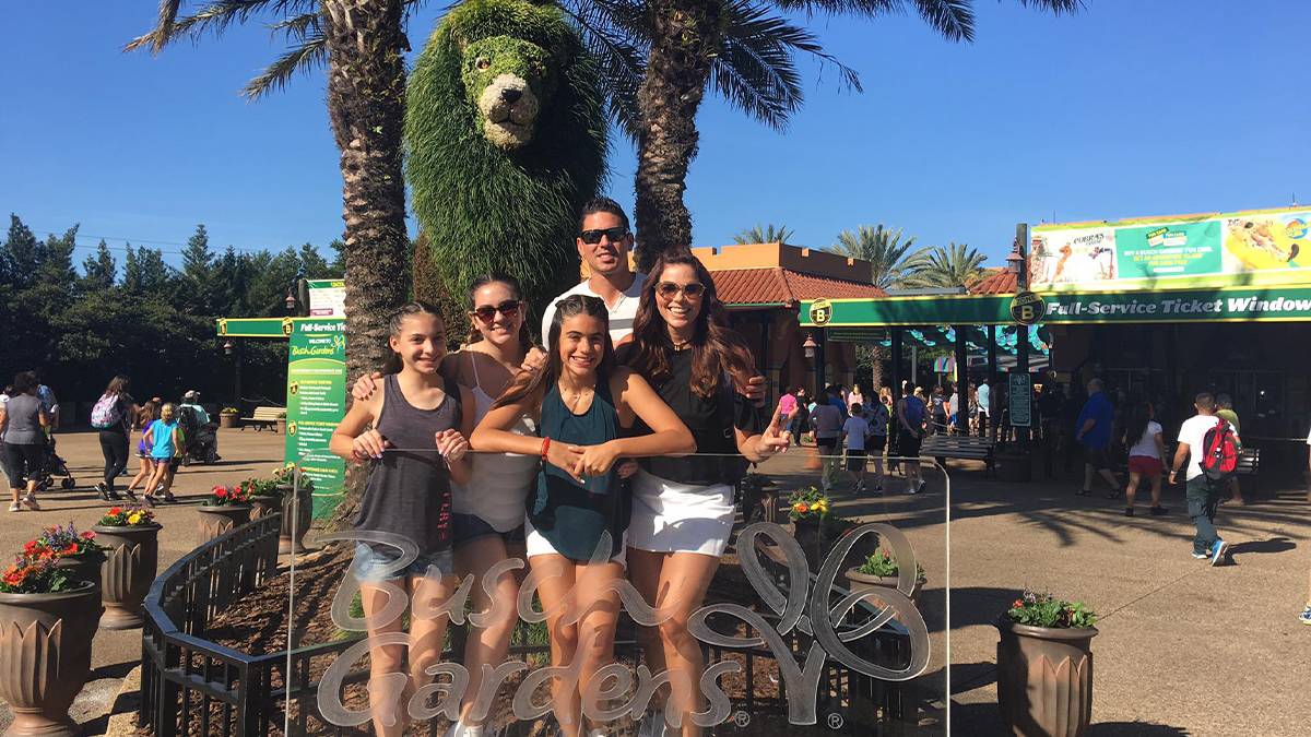 A family wearing tank tops posing for a photo behind a glass Busch Gardens Tampa sign and a lion shaped shrub behind them in Tampa, Florida, USA